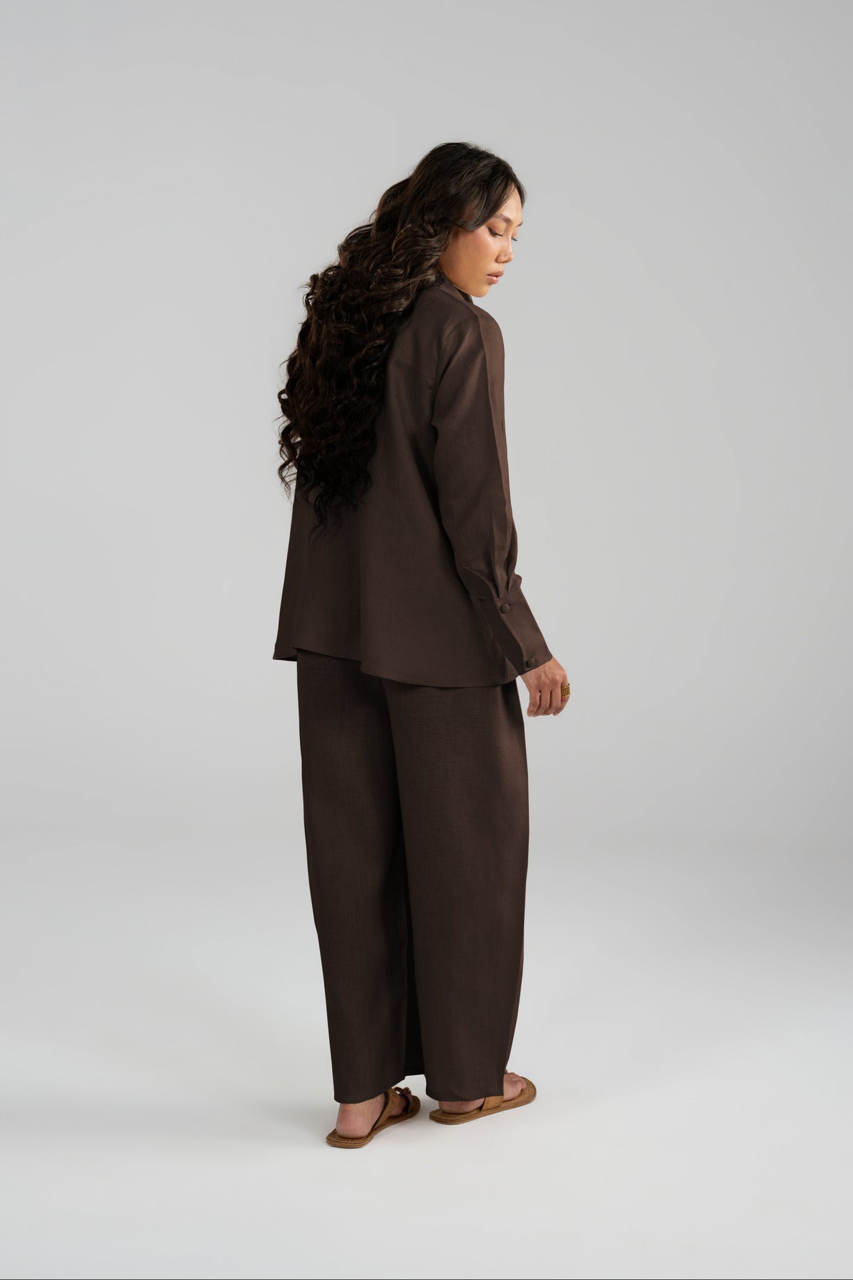 A woman with long, wavy dark hair is shown from a side-back angle wearing a matching dark brown two-piece linen set consisting of a long-sleeve top and wide-leg trousers.