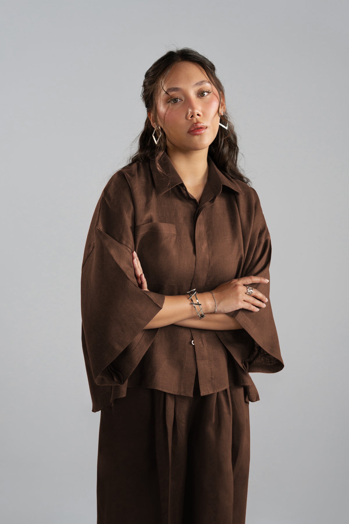 Model posing in a dark brown two-piece linen set with broad collars, loose wide sleeves and silver jewelry.