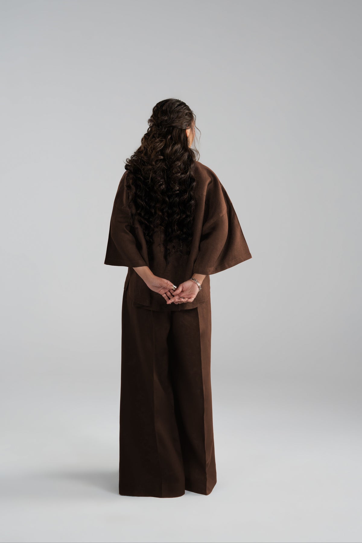 A model viewed from behind wearing a dark brown wide-sleeved tunic top and matching wide-leg trousers.