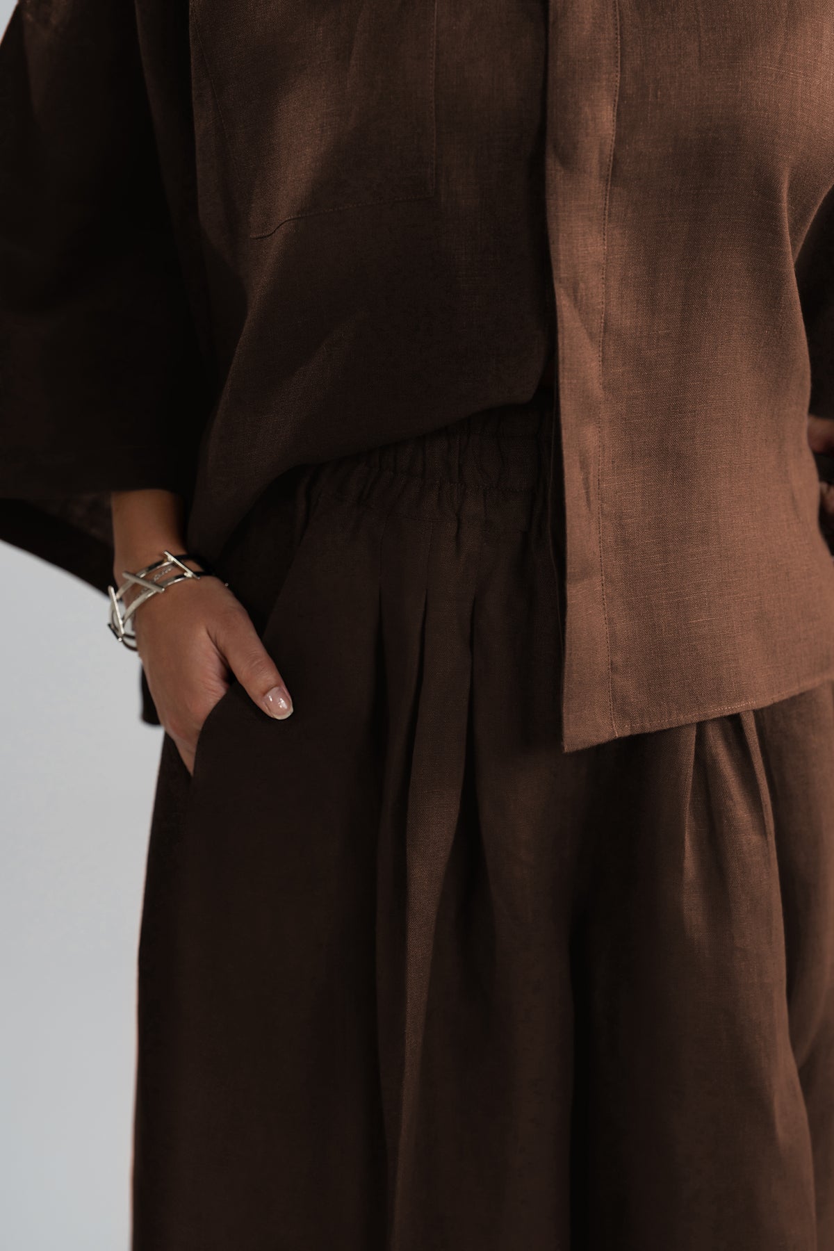 A close-up view of a dark brown linen outfit, featuring a cropped long-sleeve top and matching pleated trousers, with a silver-toned bracelet.