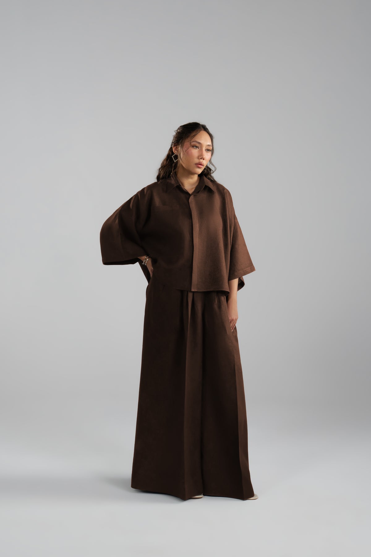 A model wearing a dark brown two-piece outfit: a cropped button-up shirt and matching wide-leg trousers.