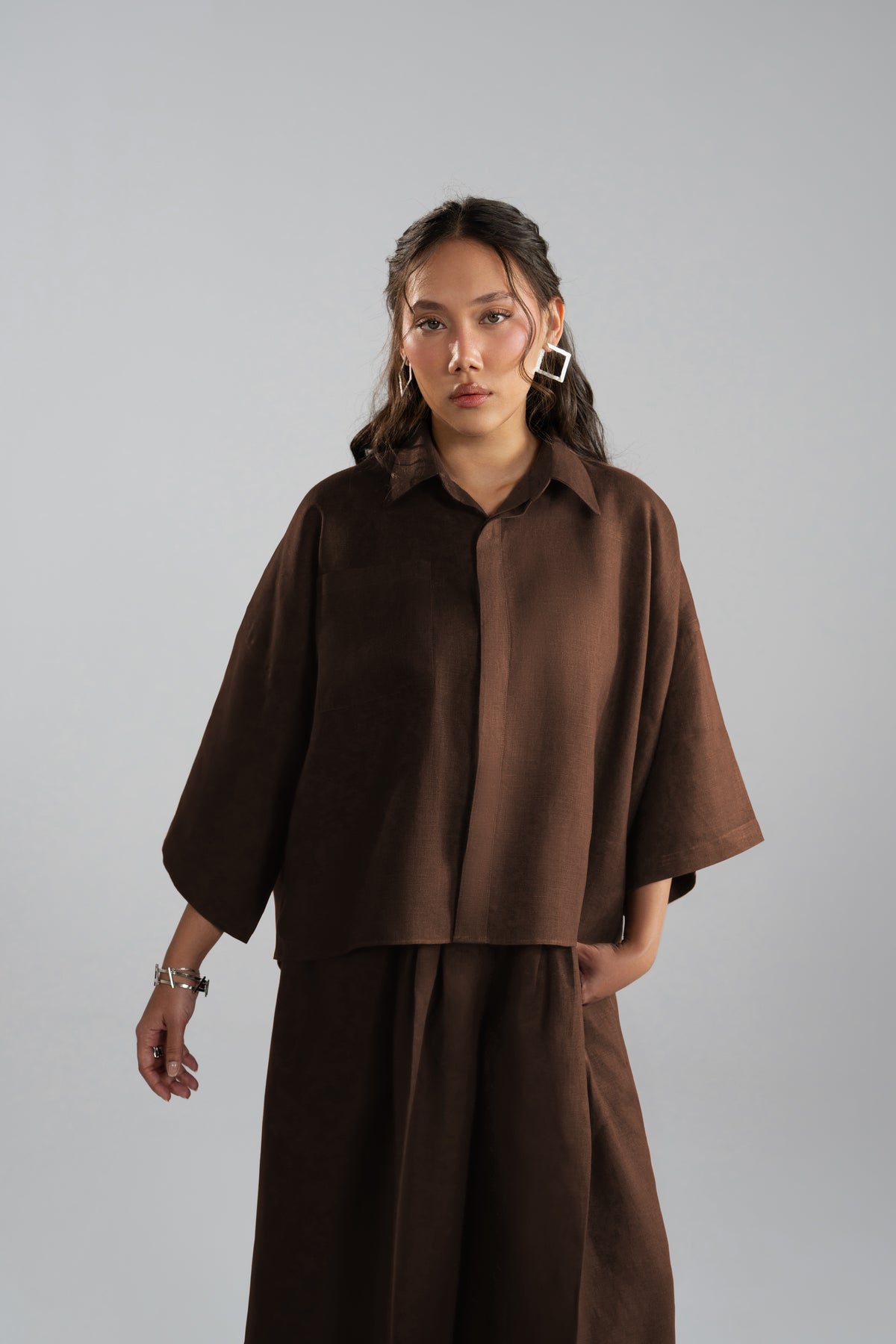 A woman modeling a dark brown two-piece outfit: a short-sleeved button-up shirt and matching wide-leg trousers.