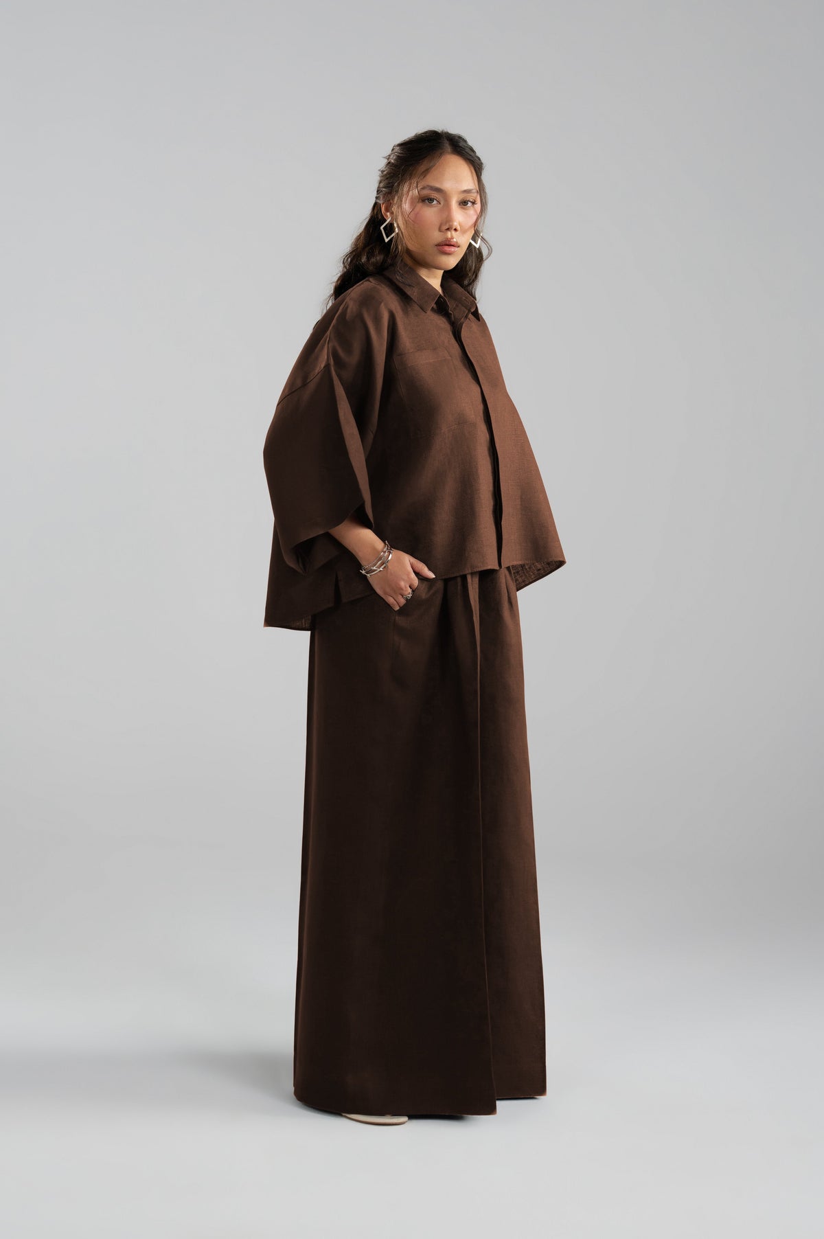 A model wearing a dark brown, minimalist two-piece outfit of pure linen consisting of a cropped long-sleeve top and matching wide-leg trousers.
