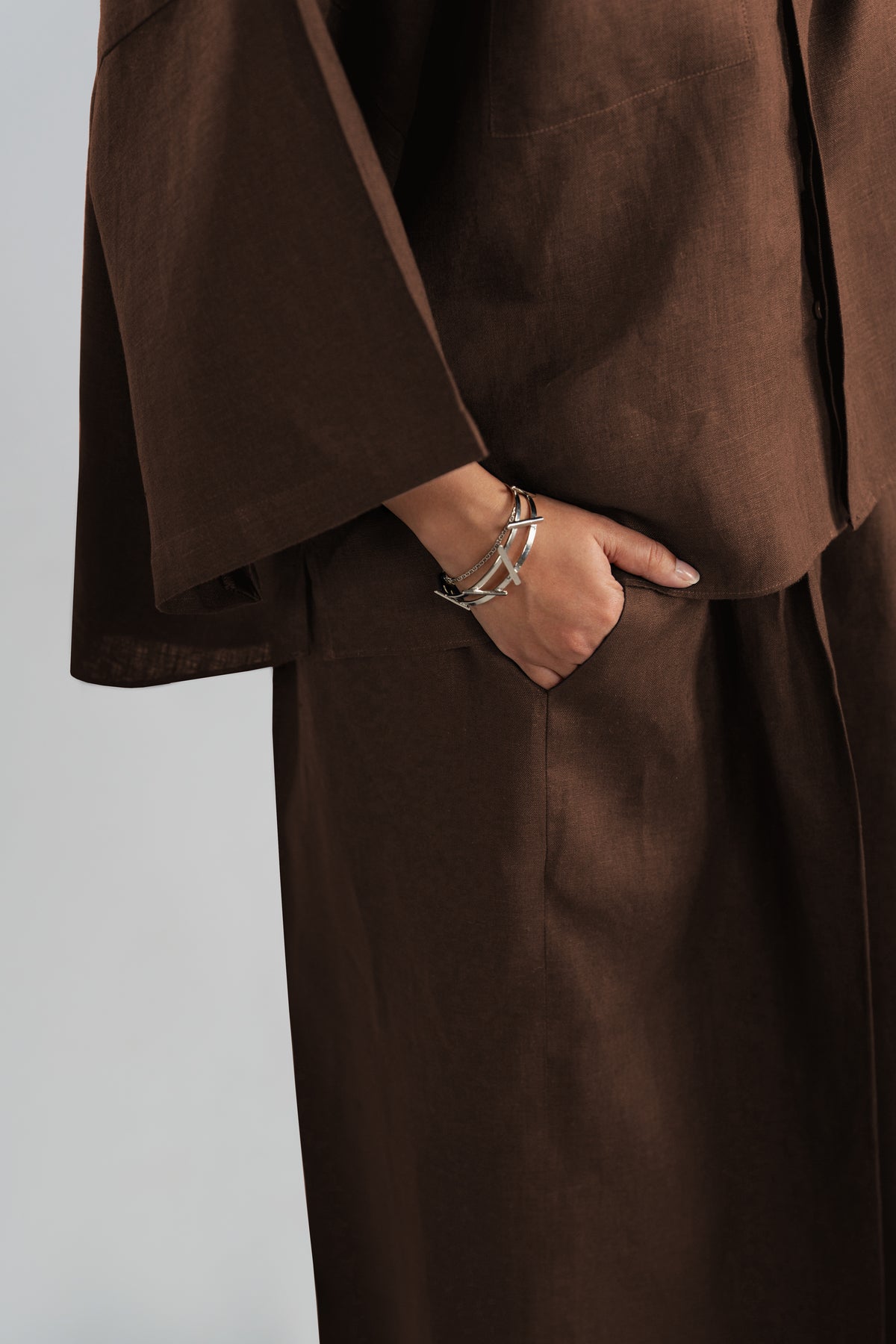 A close-up view of a pure linen dark brown wide-sleeved shirt with a matching trousers, showing one hand in a pocket and wearing silver bracelet.