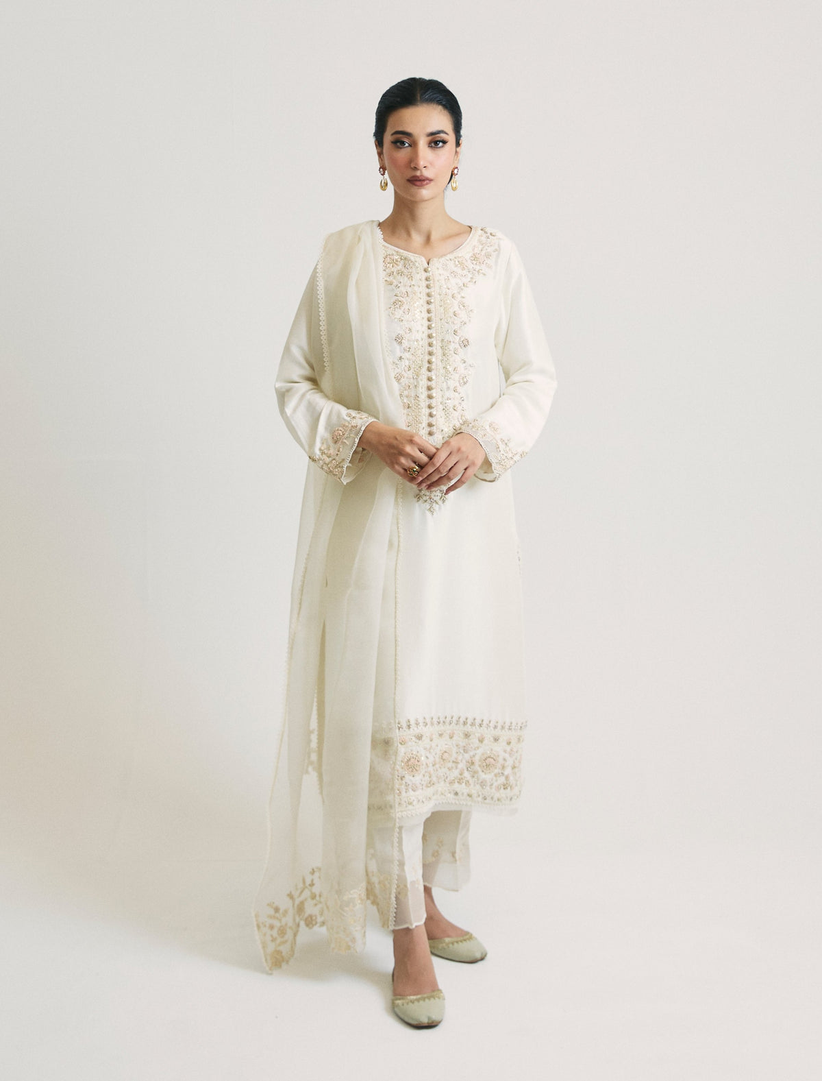 Ivory Pakistani suit in pure raw silk, worn with an organza dupatta draped over one shoulder, featuring exquisite hand-embroidered floral detailing around the neckline and along the hem border.