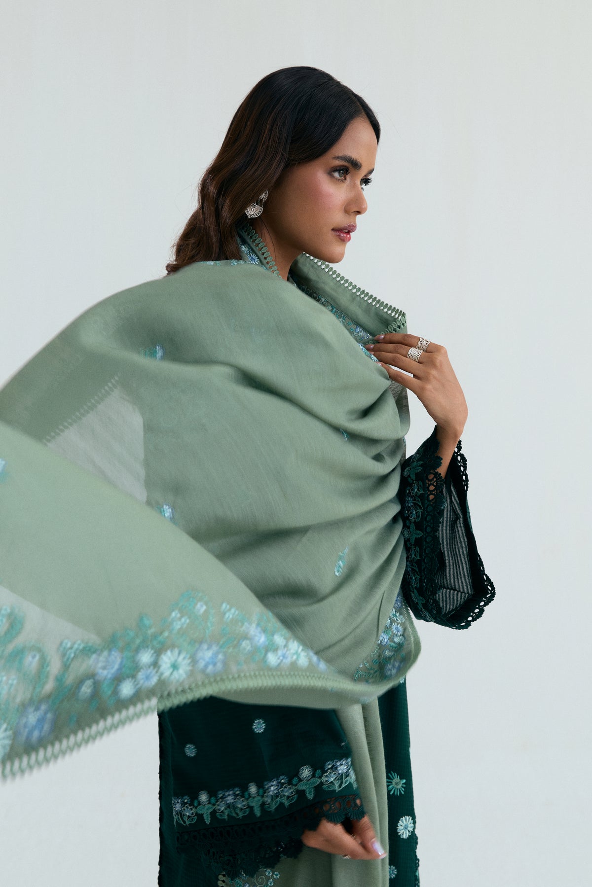 A sage green draped shawl adorned with floral border embroidery, allowing glimpses of the dark green kameez and the lace detailing on the sleeves.