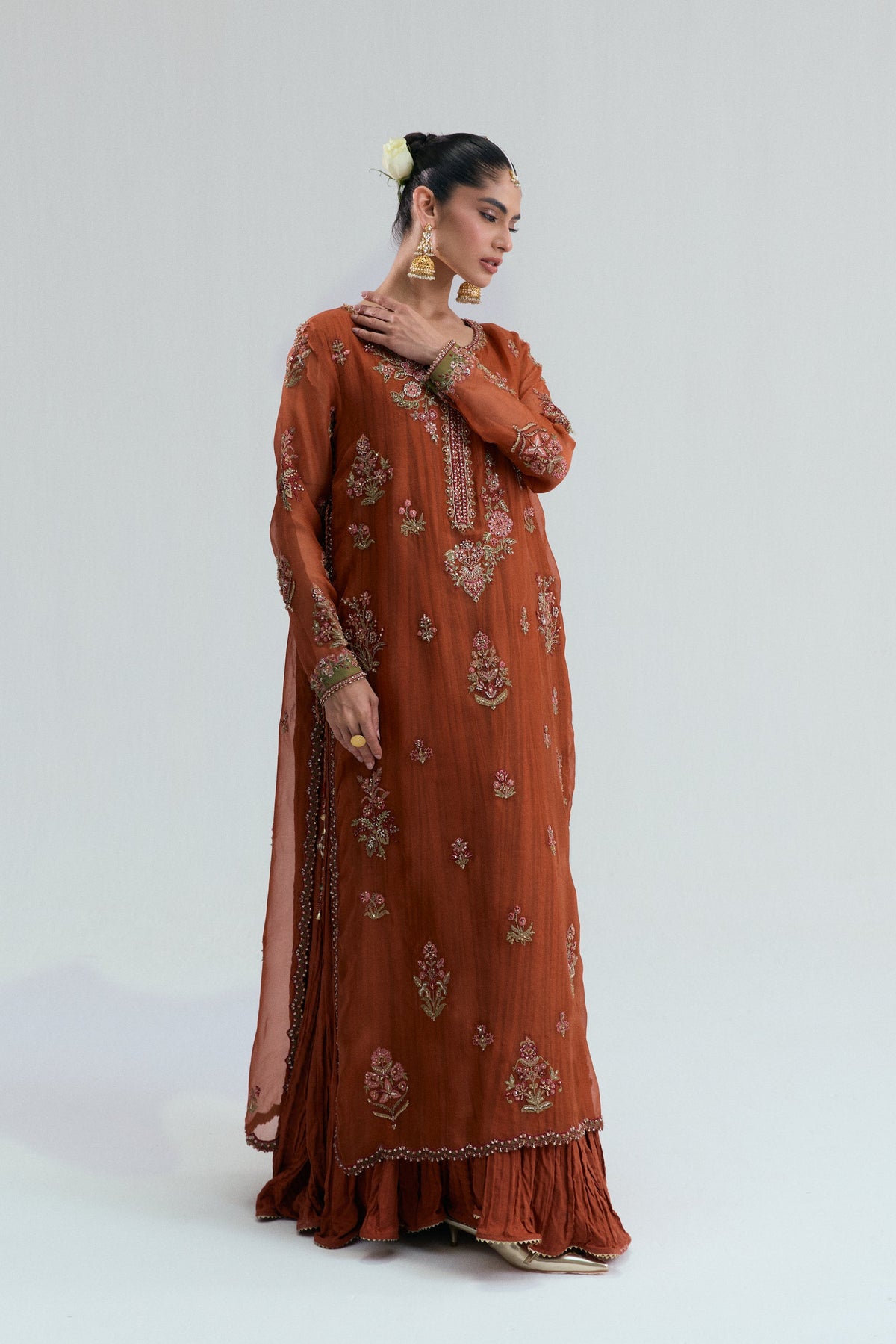 A rust-colored Pakistani women's suit featuring floral embroidery and sequins on a long V-neck kameez, paired with matching crushed, flared bottoms.