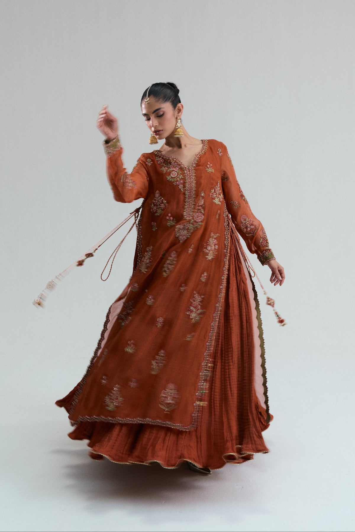 Women's rust long kameez featuring floral V-neck embroidery, side tassels, and a scalloped organza dupatta paired with a wide crushed skirt.