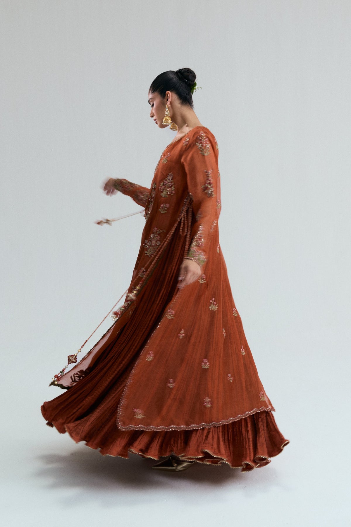 A model twirling in a rust-orange suit, showing long embroidered sleeves, a flared crushed skirt, and dupatta tassels in motion.