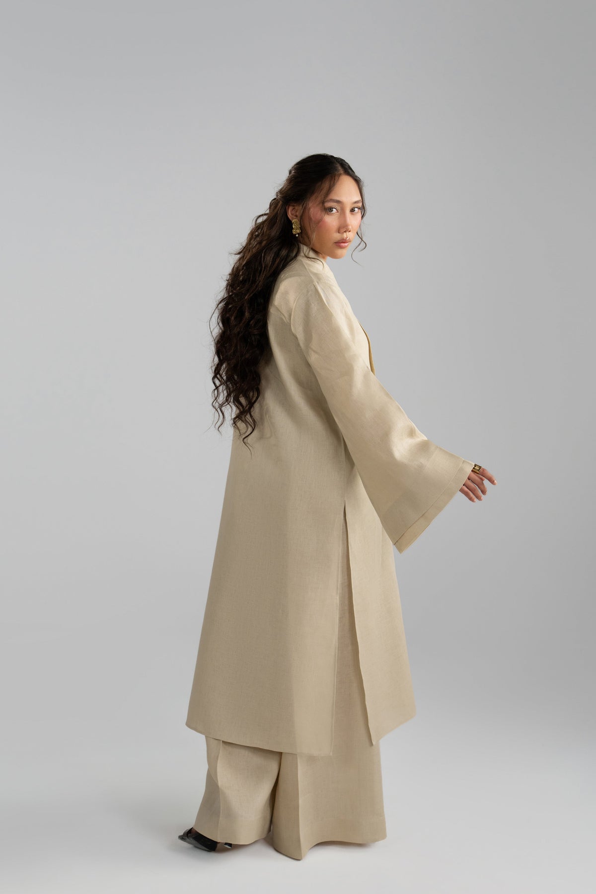 Back three-quarter view of the beige linen co-ord, showing the centre-seam tunic and bell sleeve drape