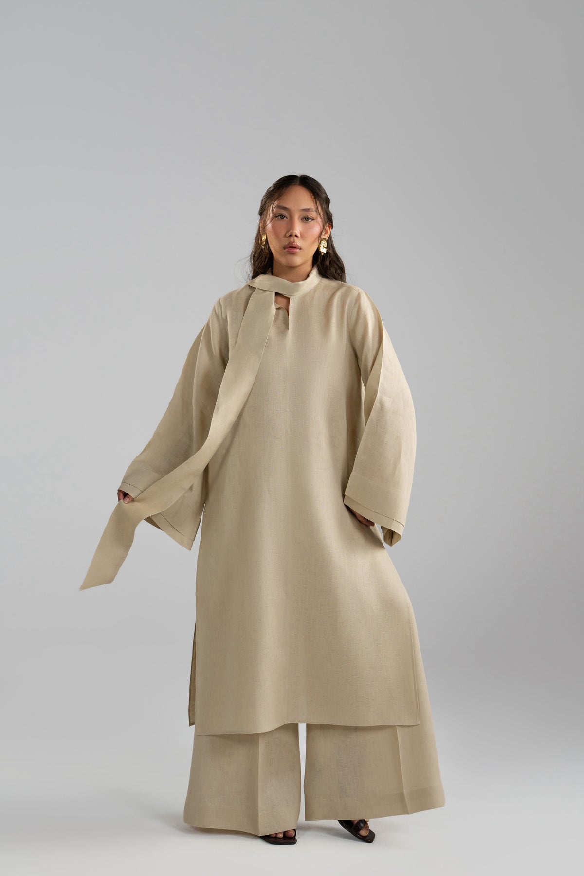 The model is holding the long scarf attached to this beige, light-brown linen summer set, paired with wide-legged, flowing trousers.