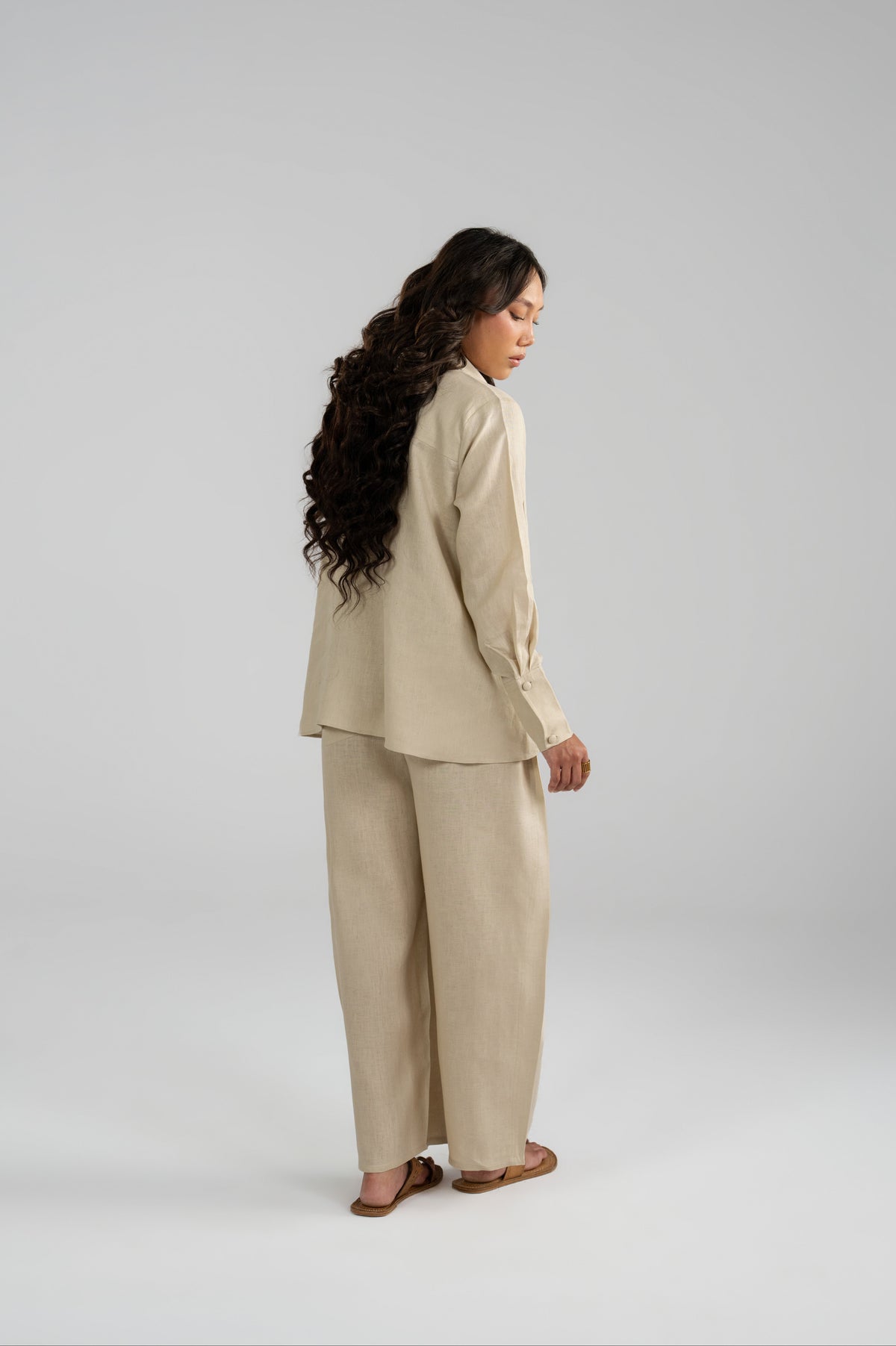 Full-length back and side profile of a model wearing the Tuscany beige pure linen set, showing the rear drape of the short shirt and wide balloon trousers.