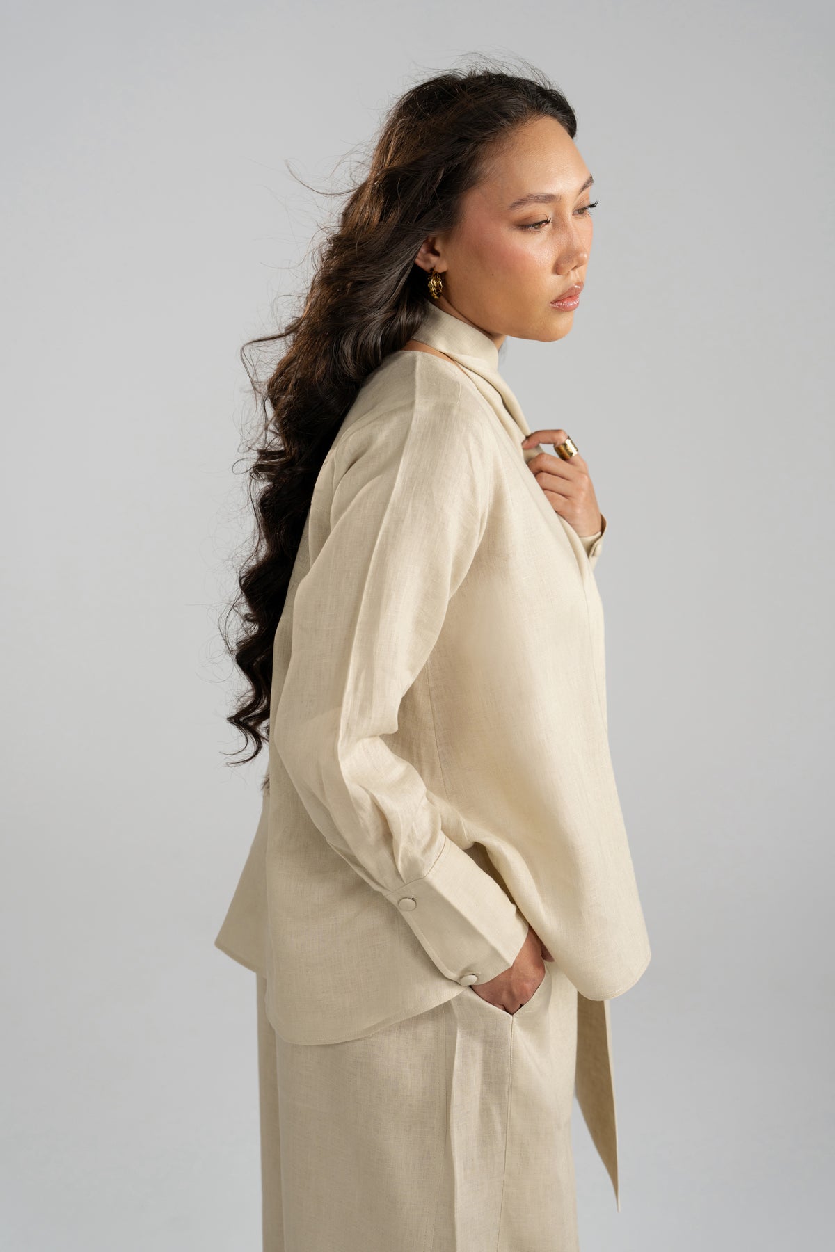 Three-quarter side profile of Tuscany beige pure linen top, highlighting long gathered sleeves with button cuffs and hand in pocket.